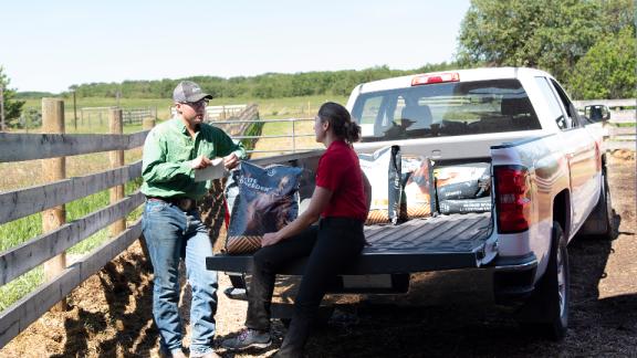 A woman sitting in the bed of a truck with bags of feed, talking to a farmer in the feed yard